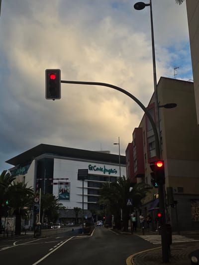 The image depicts a street scene in Santa Cruz de Tenerife, Spain. In the foreground, a wet asphalt road with white lane markings curves to the right. A pedestrian crossing is visible on the right side of the road, marked with white stripes. A traffic light displaying a red signal is positioned above the road.
In the middle ground, several palm trees line the sidewalk. Behind them, a large, modern building with the logo "El Corte Inglés" is prominent. The building has a sleek, white facade with dark-tinted windows and a prominent angled roofline. To the right of El Corte Inglés, a multi-story building with red and yellow sections is visible. Several people are standing on the sidewalk near the pedestrian crossing, some appearing to wait. Another traffic light, also showing red, is located on the right side of the image, with a pedestrian signal displaying a green walking figure.
The background is dominated by a cloudy sky, with patches of yellow and pale blue suggesting the presence of the sun behind the clouds. A street lamp stands tall on the right side of the frame. The overall impression is a street scene in an urban environment during daylight hours, possibly after rain.