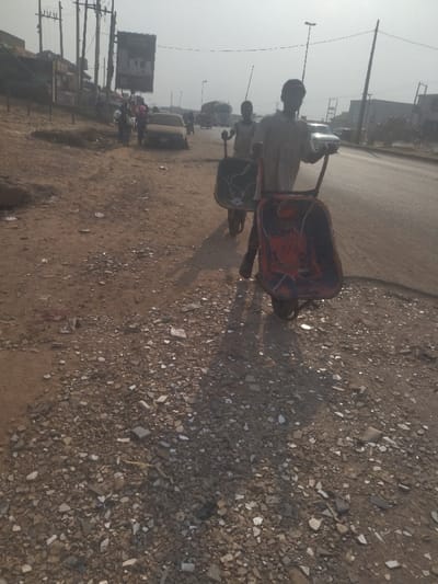 The image shows two young men pushing wheelbarrows along a dirt road in Way, Nigeria. The road is covered with gravel and rubble. There is a car parked in the background. The men are wearing casual clothing. The wheelbarrows are both empty. One wheelbarrow is red with black and white markings, and the other is black. The scene is captured during the daytime.