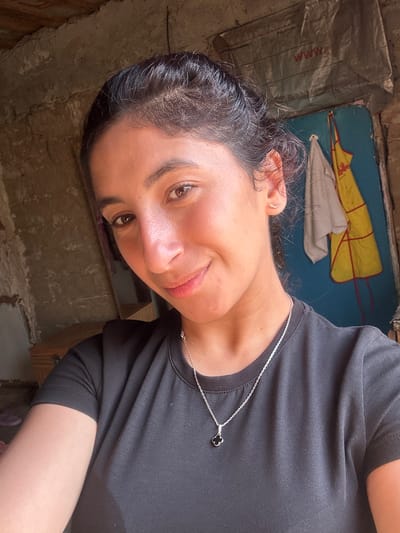 A young woman with dark hair pulled back in a bun, is looking directly at the camera with a slight smile. She is wearing a black t-shirt and a silver necklace with a clover-shaped pendant. Her left ear is adorned with a small, curved earring. The background appears to be an indoor setting with rough, textured walls. To her right, a blue surface is visible with a yellow and white garment hanging on it. The overall lighting suggests a natural light source from the left. This image does not contain any elements that would place it in the city of Dondukovskaya, Russia.