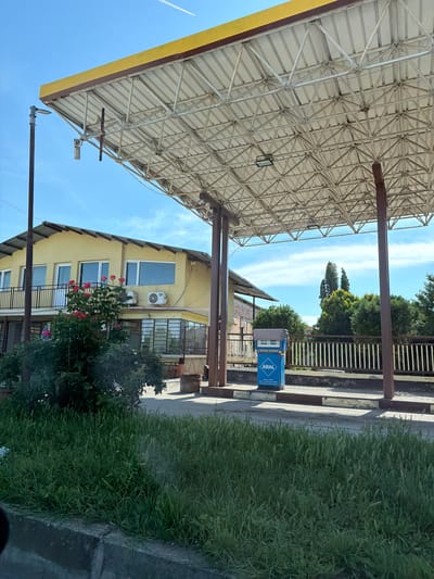 This media file captures an outdoor scene, likely at a gas station or a similar commercial facility in a suburban or rural setting, given the context of Brestovets, Bulgaria. The primary view is from under a large, light-colored overhead canopy, which is supported by a complex metal truss framework and several sturdy, dark brown pillars. The canopy features a yellow trim along its edge, and a light fixture hangs from its underside.
In the mid-ground, a distinct blue and white fuel dispenser is visible, prominently displaying the brand name "ARAL." Below the brand name, smaller text can be partially deciphered, including "Main function" and "For optimal fuel...". Behind the dispenser, a light-colored metal fence or railing separates the paved area from a line of various green trees.
Further back, a multi-story building with a light yellow exterior is seen. It has several windows, some with white frames, and a balcony on the lower level. An air conditioning unit is affixed to its facade. In front of the building, a bush adorned with vibrant red flowers, possibly roses, adds a splash of color.
The background features a clear, bright blue sky with scattered wispy white clouds, and a faint airplane contrail is visible. The ground consists of a paved area under the canopy, transitioning into a dense patch of tall, green grass along the right side of the frame and in the foreground.
The scene appears to be captured during broad daylight on a sunny day. No human activity or specific event is taking place, presenting a still and peaceful environment.
