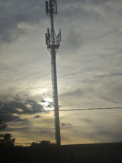 A silhouetted cell tower, approximately 50 meters tall, stands against a cloudy sky at dusk. The tower has multiple antennas mounted at its top and along its shaft. In the foreground, the dark silhouettes of trees and a building are visible. The sun is low in the sky, creating a hazy glow and casting rays through the clouds. The scene appears to be located in a rural or suburban area, possibly on the outskirts of Chepelyovo, Russia. There are no people visible in the image. The overall atmosphere is one of transition from day to night, with a slightly ominous feel due to the dark silhouettes and dramatic sky.