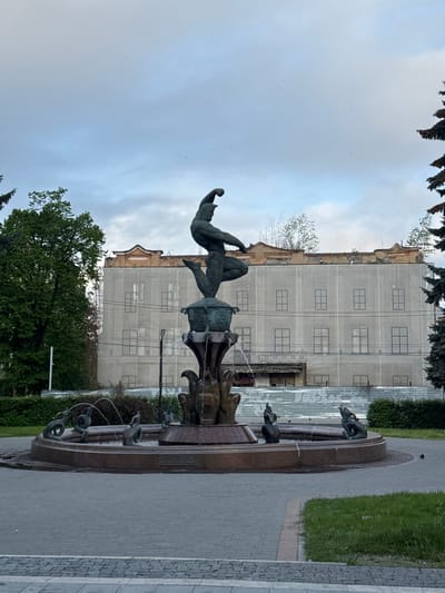 The image captures a view of the Mikhail Avakyants Fountain in Akhieli, Georgia. The fountain features a bronze sculpture of two intertwined figures, one seemingly airborne and the other grounded. The fountain itself has multiple levels and spouts water from the mouths of animal figures into a circular basin. Behind the fountain is a building undergoing renovation, covered in netting with outlines of windows. To the sides are paved walkways and grass areas, with trees and bushes adding greenery to the scene. The sky is overcast, suggesting a cloudy day.