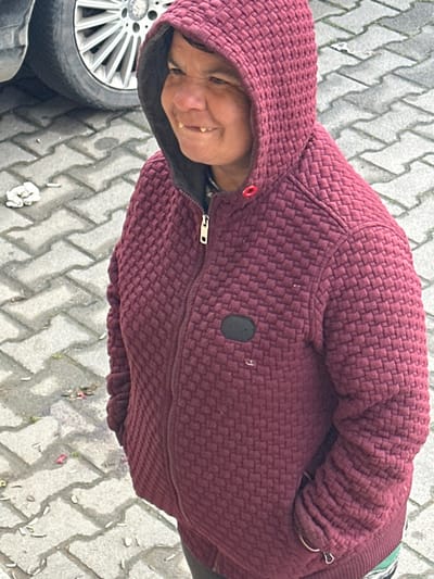 An individual is depicted in a medium shot, standing outdoors in Plodovitovo, Bulgaria. The subject, an adult with medium skin tone, wears a dark red/maroon hooded jacket with a textured, possibly quilted, pattern. The hood is up. The jacket is fastened with a silver zipper, partially unzipped, and features a dark oval patch on the left chest area. A red loop is visible near the collar. The individual's head is turned slightly to the left, with eyes directed towards the left side of the frame. A smile or grimace is present, exposing multiple discolored upper and lower teeth, including a noticeable gap between the two upper central incisors. The background consists of a grey interlocking concrete paver surface laid in a herringbone pattern. In the upper left, a dark vehicle tire and a silver multi-spoke wheel rim, consistent with a Mercedes-Benz, are partially visible. Scattered debris on the pavement includes crumpled white items, several cigarette butts, and small pieces of dark plant matter.