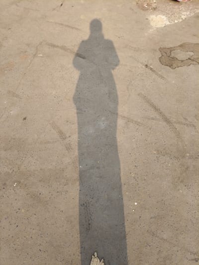 The image displays the elongated shadow of an individual cast upon a light gray, granular paved surface, likely concrete or asphalt, in Bardhaman, India. The shadow, extending vertically from the upper-center to the bottom-center, portrays a human silhouette with apparent folded arms or an object held in front. Multiple faint, dark tire tracks are visible across the pavement, notably to the right and intersecting the shadow's mid-section. An irregular, dark liquid stain is present in the upper right quadrant. Fine, linear cracks traverse the ground in various locations. A small, light-colored piece of debris is partially obscured by the shadow at the bottom edge. The extended shadow indicates a low sun angle.