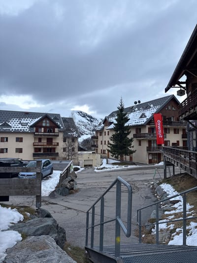 The image displays an alpine village street scene in Saint-Sorlin-d'Arves, France, under an overcast, grey sky. The environment indicates a winter or early spring setting, with snow patches present on the ground and building rooftops.
In the immediate foreground, on the right, a set of grey metal grate stairs with a corresponding metal handrail descends out of view. A yellow mark is visible on the right-side railing. To the left, large grey boulders and sparse, dry grass are interspersed with remnants of snow.
The midground is dominated by a paved road that curves through the village. On the left, a wooden fence borders a parking area containing a light blue Volkswagen Golf, partially obscured by a dark vehicle, both parked on snow-dusted ground. Behind this, a multi-story building with a light-colored facade, brown window frames, and wooden balconies features a dark grey roof with snow accumulation. Centrally, a tall, slender evergreen tree stands prominently. To its right, another multi-story building, exhibiting a light-colored facade and wooden upper sections, displays a vertical red banner with white, reversed text. This building also has multiple balconies and a snow-covered dark roof, with a wooden elevated walkway extending from its right side. Further along the curving road, a smaller, light-colored building with a closed garage door is visible.
The background features rugged, snow-covered mountains under the consistently heavy, grey cloud cover. No individuals or active interactions are depicted within the scene.