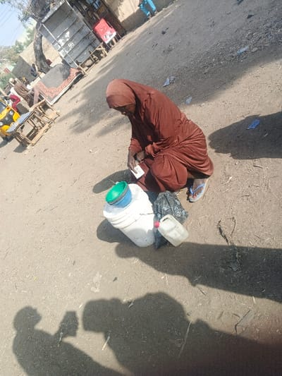 The image depicts an outdoor scene in Gobirawa, Nigeria, centered on an elderly individual, likely female, squatting on unpaved dirt ground. This individual wears a brown head covering and a matching loose-fitting brown garment, with blue flip-flops visible on their feet. Their hands are positioned over a small, light-colored rectangular object. Adjacent to the individual, on the ground, are a white plastic bucket with a green lid and a blue bowl stacked on top, a crumpled black plastic bag, and a translucent white plastic jerrycan with a red cap. Multiple distinct shadows are cast on the ground. In the mid-ground, a yellow and black three-wheeled vehicle is partially visible, along with various wooden market stalls or shelving units, including a large, empty white-painted structure. A patterned rug is leaned against one of these structures. Other individuals in traditional attire and parts of buildings with light-colored walls are visible in the background. The ground is uneven dirt, strewn with small pieces of debris. The scene suggests a busy market or street area under direct sunlight.