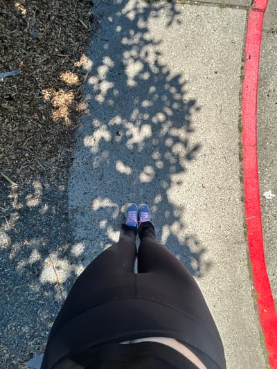The image shows a person's legs and feet from a first-person perspective. They are wearing black leggings and purple sneakers. The person is standing on a paved sidewalk with a red curb to their right. To their left is an area covered in wood chips and gravel. The ground is dappled with shadows from tree leaves. The scene appears to be outdoors in a city, likely Everett, United States of America, given the context.