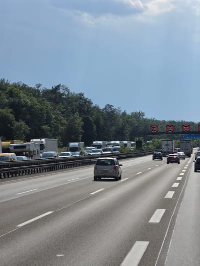 This image depicts a traffic jam on a highway in Neu-Isenburg, Germany. The scene is set on a multi-lane road, with a dense line of cars and trucks stretching into the distance. The vehicles are primarily modern, including several trucks with trailers, passenger cars, and vans. The road is paved with asphalt and marked with white lane dividers. On the left side of the highway, a dense forest of green trees lines the road, suggesting a rural or suburban setting.

Several digital speed limit signs are visible, all displaying "120". These signs are mounted on gantries over the highway. The sky is a clear, light blue with a few wispy white clouds, indicating fair weather. The sun appears to be shining, casting shadows and illuminating the scene, suggesting it is daytime, likely mid-morning or afternoon.

The primary activity is the standstill traffic, with vehicles stopped or moving very slowly. The overall mood conveyed is one of frustration or patience, typical of a traffic congestion. There is a visible license plate on one of the cars in the foreground, reading "43-KK-PD". The perspective is from a slightly elevated position, looking down the highway.
