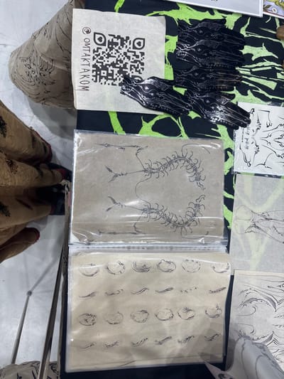 The image depicts an overhead view of a display table in Ban Rong Ruea Kham, Thailand, covered with a black fabric featuring a neon green abstract pattern. On the table, various merchandise is arranged. In the upper left quadrant, a square piece of light brown paper exhibits a black QR code above the text "@OMTAKTAKOM". Adjacent to this, multiple black, hand-shaped objects with intricate silver wavy patterns are stacked and spread. Below these, two clear plastic sleeves contain sheets of light brown paper. The top sleeved sheet displays a black line drawing of a segmented, centipede-like creature forming a circular or heart-shaped motif, flanked by spear-like appendages. The bottom sleeved sheet presents multiple rows of smaller, distinct black line drawings, including wavy lines and irregular shell-like or organic shapes. To the right, additional sheets of paper with black line art, some resembling skeletal or anatomical structures and abstract wave patterns, are visible. On the far left, the lower body of an individual is partially visible, wearing a patterned light-colored garment and red footwear. A thin, silver-colored metallic pole is visible in the lower left foreground, and a light-colored floor extends beneath the display.
