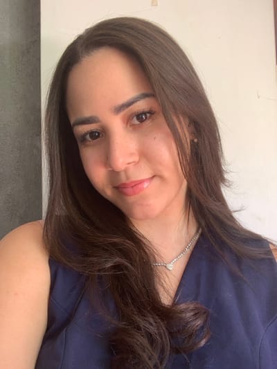 A frontal portrait photograph captures a single female individual in an indoor setting located in Lauro de Freitas, Brazil. The subject, an adult with a fair complexion, brown eyes, and long, dark brown hair parted on the left, is positioned facing the viewer with a slight smile. She is wearing a dark blue, sleeveless garment and a silver-colored chain necklace featuring a small heart-shaped pendant. A light-colored earring is partially visible on her right ear. The background on the left consists of a dark gray, textured wall, while the background on the right is a plain, light-colored wall that shows a small, light-brown rectangular mark in the upper right quadrant. The scene is uniformly illuminated.