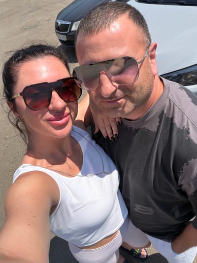 Two people, a man and a woman, are posing for a selfie outdoors. The woman is on the left, wearing a white crop top and sunglasses. The man is on the right, wearing a grey t-shirt and sunglasses, with his arm around the woman. They are standing in front of a white car. The ground is paved with asphalt. In the background, a dark car is visible. The scene appears to be in a parking lot.