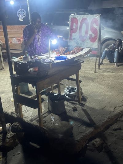 The image depicts a nighttime scene in Abraka, Nigeria, featuring a street vendor operating a roadside grill. The vendor, a man wearing a purple patterned shirt, is seen on the left side of the frame, seemingly on a phone call. He stands behind a makeshift wooden table displaying grilled meat. A POS (Point of Sale) sign is visible above the table. A white car is partially visible in the background, and a second person can be seen near the car. Debris and litter are scattered around the base of the grill stand and on the pavement. The street is dimly lit, with shadows cast by the grill and the table. The ground is paved with interlocking concrete blocks.