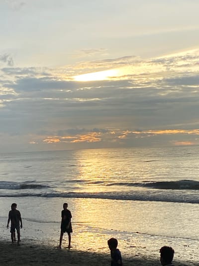 The image depicts a beach scene at sunset in Bateetimoh, Indonesia. The sky is filled with clouds that are illuminated by the setting sun, creating a warm, golden glow. The sun is partially visible through a gap in the clouds, casting a bright reflection on the calm ocean water. Gentle waves lap at the shore, where several silhouetted figures are present. In the foreground, three individuals are visible on the sand, two in the mid-ground and one in the far right foreground. They are facing the ocean, some appearing to be walking or standing. The overall atmosphere is serene and tranquil, characteristic of a tropical sunset.