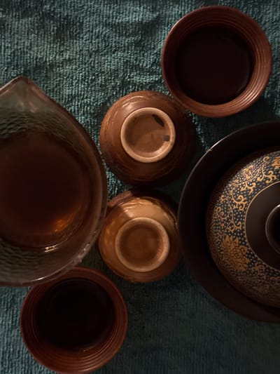This is an overhead, close-up shot of a collection of ceramic tea ware on a teal-colored, textured surface, possibly a tablecloth. From top to bottom and left to right, the items include: a large, dark brown ceramic pitcher with a hammered texture, a smaller, lighter brown ceramic bowl turned upside down, another similarly colored bowl also upside down, a large, dark serving tray with ornate gold and black patterns, and a dark brown ceramic bowl with ribbed sides. The scene appears to be a still life, possibly arranged for aesthetic purposes. There are no people visible in the image, and no actions or interactions are taking place. The environment is solely defined by the objects and the textured surface they rest on. The provided image and its crops do not contain any information that would allow for the identification of its location within the city of Pevek, Russia.