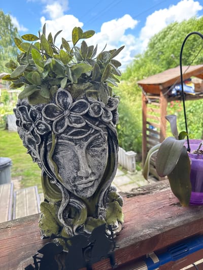The image displays a weathered, concrete planter in the shape of a woman's head, adorned with flowers and greenery, positioned on a wooden railing. The planter is filled with artificial green foliage. To the right, an orchid plant in a purple pot is visible. The background shows a lush, green garden with trees, a wooden structure with a brown roof, and a blue sky with scattered clouds. The location is identified as Yesipovo, Russia.