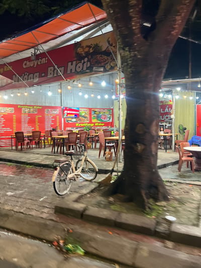 The image depicts an outdoor dining area of a restaurant, likely located in Minh Xuân Ward, Vietnam, at night. String lights illuminate the scene, casting a warm glow on rows of tables and chairs set up on a paved ground. A large tree trunk dominates the foreground, its rough bark contrasting with the man-made structures. In the middle ground, a light-colored bicycle is parked near some of the tables. Large red banners with Vietnamese text and images of food hang on the restaurant's walls, advertising menu items. The ground is wet, suggesting recent rain, and there is some debris visible along the edge of the paved area. A person is seated at a table in the background. The overall atmosphere appears to be that of a casual, open-air eatery.
