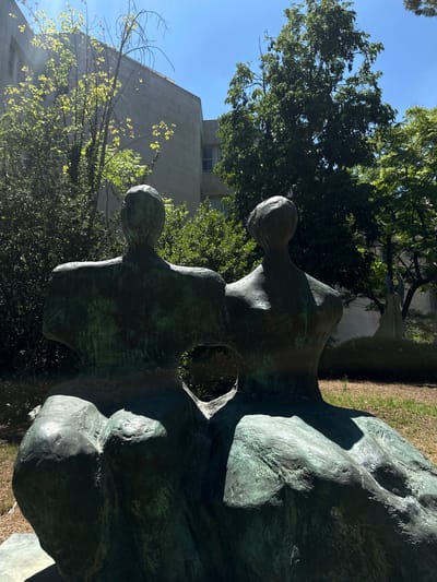 This media file shows a close-up view of a large, weathered bronze sculpture situated outdoors. The sculpture appears to be abstract or semi-figurative, with smooth, undulating surfaces and rounded forms that suggest human figures in repose or contemplation. The patina on the bronze varies, with areas of green oxidation contrasting with the darker, metallic tones. Sunlight casts strong shadows across the sculpture, highlighting its textures and contours.
In the background, lush green trees and foliage are visible, along with a portion of a light-colored, modern building. The sky above is a clear, bright blue, indicating a sunny day. The overall impression is one of serene contemplation and the interplay of natural light with man-made art and architecture. The setting suggests a park, a public garden, or the grounds of an institution. There is no visible text in the image.