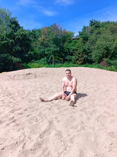 A middle-aged man with short, dark hair and a tattoo on his left arm sits on a sandy embankment. He is shirtless, wearing dark swimming trunks, and his feet are covered in sand. The embankment is part of a beach, with the sea or a large body of water implied by the context. Behind the man, a line of dense green trees forms a backdrop, with a bright blue sky with wispy white clouds overhead. A volleyball net is visible among the trees. The scene suggests a warm, sunny day at a recreational area, potentially within or near the village of Vyazovka, Russia, given the geographical context.