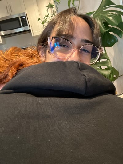 A person in a black hoodie and eyeglasses poses with shoulder-length brown hair. A houseplant with large green leaves appears in the background, slightly out of focus. A kitchen setting is visible on the left, featuring white cabinets and a stainless steel microwave. The image is vertically oriented and the camera appears to be angled downward.