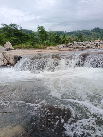 This image depicts a natural river environment in the vicinity of Meurah, Indonesia. In the foreground, turbulent river water flows towards the viewer, exhibiting extensive white aeration, foam, and small bubbles, particularly concentrated in the lower half of the frame. A low, grey concrete or stone weir extends horizontally across the midground, with water actively cascading over it, creating a visible sheet of falling water, spray, and white foam as it plunges into the downstream section.
To the immediate left of the weir, large, irregular grey boulders are visible on the riverbank. To the right, the riverbank consists of a dense collection of smooth, rounded river stones and rocks of various sizes and shades, predominantly grey and light brown. These stones extend into the midground. Beyond the river, the background reveals a dense, verdant forest covering undulating hills, characterized by a variety of trees including broadleaf species and some palm trees. The sky above is uniformly overcast with grey clouds. No human figures or evidence of human activity are visible within the scene.