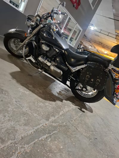 A black cruiser motorcycle is parked on a concrete floor in what appears to be an indoor parking garage. The motorcycle is angled, with the front wheel on the left and the rear wheel on the right. It has chrome accents on the engine, handlebars, and wheels, which are spoked. Black saddlebags are attached to the rear of the motorcycle. The background shows a grey wall with windows and some yellow markings on the floor, characteristic of a parking area. The lighting suggests an artificial light source. No people are present in the image. The image is a medium shot, capturing the motorcycle from a slightly low angle. The overall scene is industrial and utilitarian. The location is identified as Barri De Porta, Spain, though this specific detail is not visually discernible from the image itself.