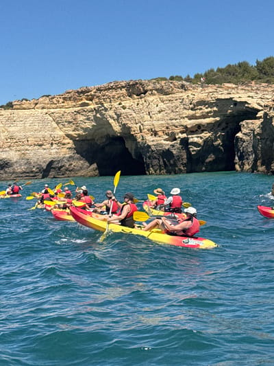 A group of kayakers, wearing life vests, paddle in the blue ocean water near a rocky cliff face with an opening resembling a cave. The kayaks are primarily yellow and red. The sky is clear and blue. The cliffs are tan and textured, with some vegetation growing on top. The water is choppy.