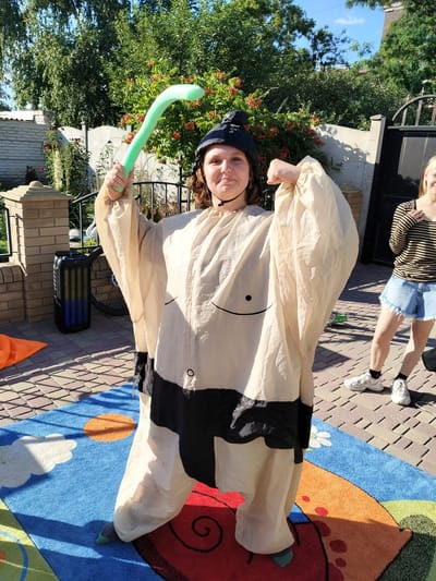 In the city of Staryi Aidar, Ukraine, a young individual, possibly female, stands on a colorful rug outdoors. They are wearing a costume that resembles a sumo wrestler's outfit, which is cream-colored with black bands. The individual also has a black cap on their head and is holding a curved, light green inflatable object in their right hand. Their left arm is flexed, showcasing their bicep. Behind the main subject, another individual, likely female, is partially visible, wearing a striped shirt and denim shorts. The background features a fence, greenery, and a brick wall, suggesting a residential or yard setting. The ground is paved with interlocking bricks, and a large speaker is visible to the left of the main subject. The lighting suggests it is daytime.