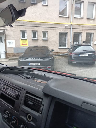 The media file captures an outdoor scene, likely a parking area or street in Svémyslice, Czechia. The viewpoint is from inside a vehicle, looking through the windshield.
The primary subjects are two cars. The car closer to the viewer, on the left, is a dark-colored Porsche Panamera. Its license plate is partially visible, showing "ELC 209K". The second car, on the right, is a dark-colored Skoda Superb Combi. Its license plate clearly reads "5L7 8381". Both cars appear to be parked.
The background consists of a building with a textured, light-colored facade. Several windows are visible on the building, some with bars. A yellow pipe runs horizontally along the building's exterior. A white sign with text is mounted on the building to the left of the Porsche, and a yellow sign with a red arrow pointing left is located to the right of the white sign.
The interior of the vehicle from which the photo is taken is also visible, showing the dashboard with a Sony car radio, air vents, and a mobile phone resting on the dashboard. The scene appears to be taking place during the daytime, under overcast skies, suggesting cloudy or potentially rainy weather. There are no people visible in the frame. The overall impression is a mundane, everyday moment captured from the driver's perspective.