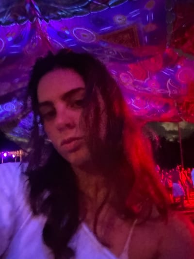 The image depicts an outdoor nighttime event in Ban Mae Ping, Thailand. In the foreground, an individual with dark, shoulder-length hair is visible, facing the camera with a neutral expression. The person is wearing a light-colored top, possibly white, with a thin strap visible on the right chest area. Their face and upper body are illuminated by purple and red artificial light.
Above the individual, a decorated fabric canopy spans the upper portion of the frame. This canopy features intricate glowing circular and geometric patterns, illuminated by purple and red lights, suggesting a festive or psychedelic ambiance.
To the far left in the background, faint purple lights indicate an unidentified structure or decorative element. To the right of the primary subject and further in the background, a crowd of multiple indistinct figures is discernible, heavily illuminated by bright red light. These figures appear to be engaged in an activity, consistent with a social gathering. Dark foliage or trees are visible above the crowd against the dark night sky. The scene is characterized by vibrant, multi-colored artificial lighting, predominantly purple and red.