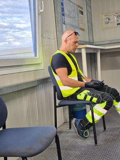 A bald man wearing black gloves, a dark t-shirt, a bright yellow safety vest, and black and neon green work pants is sitting on a dark blue office chair in a room with corrugated metal walls. He is holding a black smartphone in his gloved hands and appears to be looking at it. He has sunglasses perched on his head. The room has a large window with a white frame, showing a cloudy sky and a building in the distance. There is another empty blue office chair in the foreground. On the wall behind the man, there are several papers and maps. A blue trash can is visible under the chair. The floor is a textured metal diamond plate pattern. The man is likely on a break at a worksite. Based on the maps and signage, this location appears to be Flörsheim, Germany.