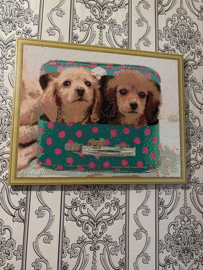 A framed diamond painting depicting two puppies in a teal box with pink polka dots hangs on a wall. The painting is made of small, colored beads that create a mosaic-like image. The wall is covered in striped wallpaper with a damask pattern in shades of gray and white. The overall scene is indoors, likely in a residential setting. There is no indication of the city of Ильпанур, Russia in the image.