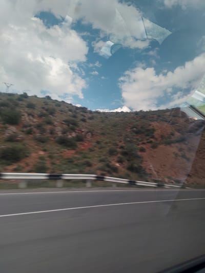 The image captures a scene viewed from inside a moving vehicle, looking out a side window. The photograph is oriented 90 degrees clockwise, causing the road to appear vertically on the right side of the frame.

On the far right, a dark asphalt road is visible, featuring two distinct white painted lines running parallel along its length. To the left of the road, a black and white striped guardrail separates it from a rugged mountainside. This mountainside, which dominates the central and left-center portions of the image, is characterized by reddish-brown soil and rocks, interspersed with sparse green and brown vegetation, primarily bushes and small trees.

The left side of the image displays a bright blue sky filled with large, white, fluffy clouds. This sky and cloud formation are also strongly reflected on the vehicle's window, particularly towards the far left, where they overlap with faint details of the vehicle's interior, possibly including part of a seat or a passenger's arm. A faint outline of a metallic lattice structure, likely a power line tower, can be seen against the sky in the lower-left quadrant.

The scene depicts a journey through a mountainous or hilly region during daylight hours under a partly cloudy sky. No discernible text is visible in the image. The location is Tigranashen, Armenia.