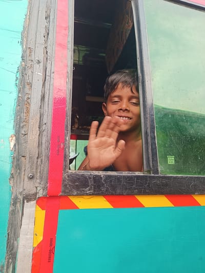 A young boy, appearing between 8-12 years old with dark, disheveled hair, is observed leaning out of a vehicle window in Kottalapalli, India. He presents a smiling expression, revealing teeth, and is shirtless. His right hand is raised and open, performing a waving gesture, with a multi-colored string bracelet visible on his wrist.
The immediate environment consists of the vehicle's window and its frame. The window frame is dark, heavily worn with peeling paint and visible fasteners. A vertical red reflective strip, marked "104R-002881" and bearing a circular emblem, runs along the left side of the frame. The closed portion of the window on the right features a green-tinted or green-colored pane, which appears textured or dirty, and displays a sticker with the text "prakash SM @K." Below the window and to its left, the vehicle's exterior is painted a prominent teal/turquoise color. A horizontal strip of yellow and red diagonal reflective markings is visible directly beneath the window. The interior of the vehicle behind the boy is dark and indistinct.