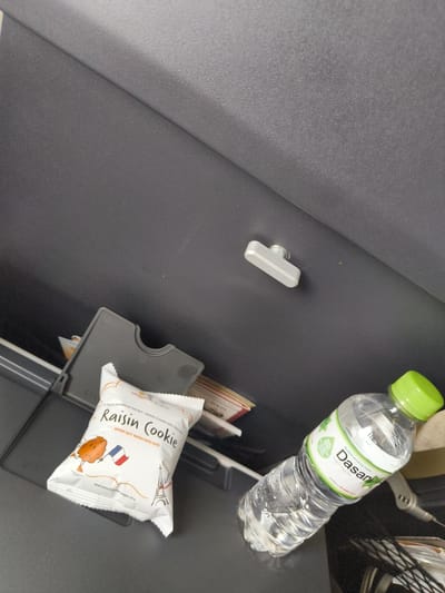 A close-up, high-angle shot captures a dark gray surface, possibly a tray table, in a seemingly enclosed space. On this surface, a small, white individually wrapped cookie package with illustrations of a cartoon cookie, the Eiffel Tower, and a French flag is prominently placed. Adjacent to the cookie is a clear plastic bottle of Dasani bottled drinking water with a green cap. To the left of the cookie package is a gray plastic holder or organizer. In the upper right quadrant of the image, a metallic silver hook-like object is attached to the dark surface. The overall scene appears to be indoors, possibly within a vehicle or airplane cabin, given the context of the items typically found on a tray table. There are no people visible in the image, and the focus is solely on the objects present.