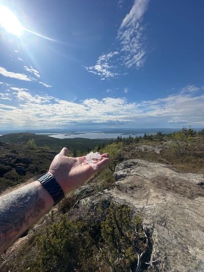 The image shows a man's arm and hand holding some small chunks of ice or frozen material. The person is wearing a black wristwatch on their left wrist. The arm is tattooed. In the background, there is a landscape featuring a body of water and wooded hills or mountains under a blue sky with scattered clouds. The scene may be from around Kandalaksha, Russia, a city located on the shore of the White Sea in the Murmansk Oblast. The environment suggests a natural, possibly elevated location overlooking the water and surrounding terrain.