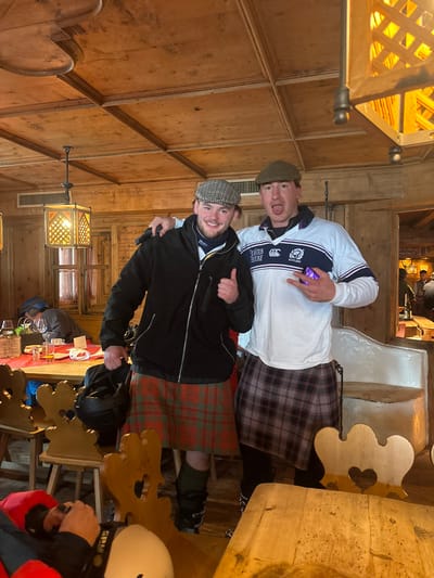 This image depicts two adult males posing indoors within a rustic establishment in St. Christoph, Austria. The environment features wood-paneled walls, a wooden plank ceiling with visible beams, and hanging wooden lanterns providing warm illumination.
The male on the left wears a plaid flat cap, a black zippered jacket over a light-colored top, and a red and green tartan kilt. He smiles at the camera, gives a thumbs-up with his right hand, and places his left arm around the shoulder of the male on the right. His lower legs are covered by dark green material, and he wears ski boots; a black ski helmet rests on a wooden chair beside him.
The male on the right wears a green flat cap, a white long-sleeved rugby shirt with blue stripes and "SCOTLAND" text, and a gray and black plaid kilt worn over dark legwear and ski boots. He poses with his tongue slightly extended and makes a "rock on" hand gesture with his right hand, while holding a small purple rectangular object in his left hand.
The foreground and midground include wooden tables and chairs featuring heart-shaped cutouts on their backs. A table on the left holds amber-colored beverages in glasses, a red napkin, and a dark green decorative item. A partially visible person wearing a dark hooded garment is seated at this table. In the background, additional indistinct figures are discernible within the establishment.