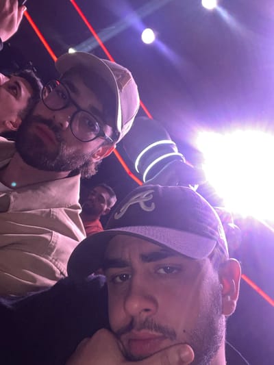 Three men take a selfie in a crowded, dimly lit venue with dramatic lighting. One man, on the left, wears a tan baseball cap, glasses, and a tan jacket. Another man, on the right, wears a dark baseball cap. A third man is partially visible behind them. The background includes bright spotlights, red neon lines, and hanging decorative elements. The lighting is stark, creating strong shadows and highlights on their faces.