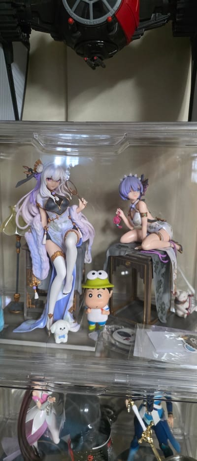 The image displays a collection of anime figurines displayed within a clear acrylic case, suggesting a retail or personal collection setting. The figurines are intricately detailed, with one featuring a woman in a light-colored, ornate dress and long, flowing purple hair, holding a fan. Beside her is another figurine of a woman with short purple hair, in a similar style dress, kneeling. A small, cartoonish character with a green hat and large eyes stands between them, along with a white dog figurine. In the background, a model of a spaceship is partially visible, hanging from above. The display case is illuminated, casting reflections on the acrylic surfaces. There is no indication of the city of Gimhae-si, South Korea, in the image itself, and no individuals are present. The scene is static and appears to be a well-organized display of collectibles.