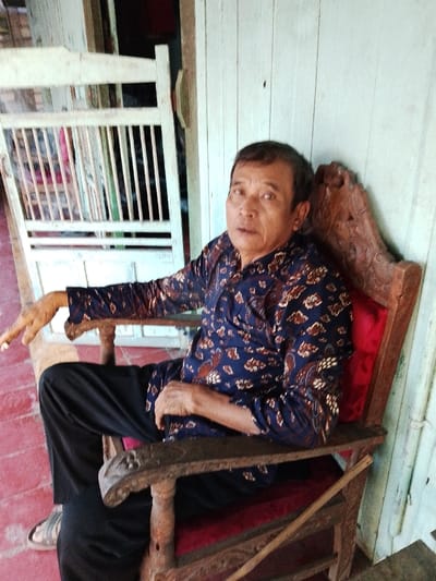 An adult male is seated in an ornate, dark-stained wooden chair with red upholstered cushions. He is wearing a dark blue long-sleeved batik shirt with brown and white patterns, black trousers, and light-colored sandals. His right arm rests on the chair's armrest with fingers slightly parted, and his left hand is positioned in his lap. The subject is looking directly at the camera with a neutral expression. Behind and to the right of the subject is a light-colored wooden plank wall, exhibiting signs of wear. To the subject's left, a partial view of a light-colored slatted wooden gate or screen is visible, with a darker interior beyond it. The floor in the foreground is reddish-brown. This scene is located in Juwana, Indonesia.