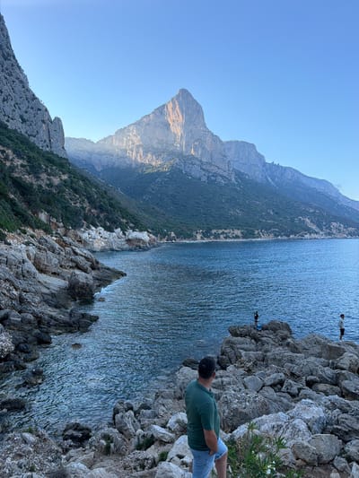 A man in a green polo shirt and denim shorts stands on a rocky shore, his back to the camera, looking out at the sea and a large, sunlit mountain. The sea is a deep blue with gentle waves. In the distance, on a rocky outcrop in the water, two other figures are visible, one appearing to be fishing. The coastline is rugged with rocky terrain and sparse vegetation leading up to imposing mountains. The sky is a clear, pale blue, suggesting it is either early morning or late afternoon. The scene depicts a tranquil coastal landscape near Baunei, Italy, possibly overlooking the Gulf of Orosei.