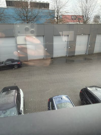 The image depicts an elevated view of a commercial or industrial outdoor scene in Apeldoorn, Netherlands. The foreground features a dark, horizontal railing. The midground consists of a wet, grey paved lot with interlocking bricks, showing visible puddles and damp areas. Three vehicles are present: a black convertible with its top down, parked on the left; a white SUV with a black roof, parked at the bottom left, facing away; and a dark blue sedan, parked at the bottom right, also facing away.

The background is dominated by a single-story building with grey paneling and five light-colored, roll-up garage doors. Reflected prominently in the glass from which the photograph was taken is a human hand holding a light-colored smartphone with a multi-lens camera system. Beyond this building, a light blue building with multiple HVAC units on its roof is visible, along with a red building displaying a sign reading "HANDS" and "VERS." Bare, deciduous trees without leaves are scattered across the upper portion of the frame, set against an overcast, light grey sky. The scene suggests a cool, damp environment.