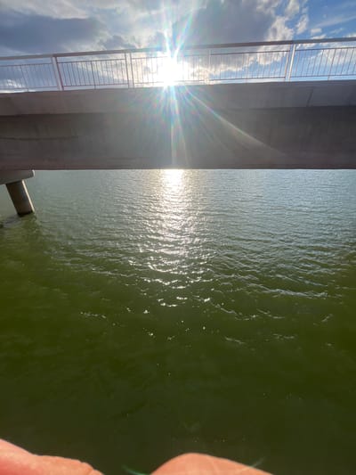 The image, taken from an elevated position in Ait Mazigh, Morocco, showcases a water body adjacent to a structure, likely a bridge or building, featuring a metal railing. The sun is positioned behind the railing, creating a strong glare and lens flare. The water surface displays sunlight reflections, with a dark green hue suggesting possible depth or algae content. A blurry object is on the right side of the water, but its nature remains undetermined. The sky, visible through the railing, is bright with scattered clouds.