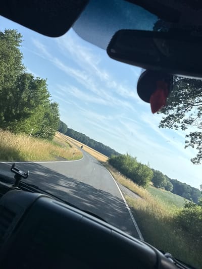 The image, taken from inside a vehicle, shows a winding asphalt road in the municipality of Oberwetz, Germany. The road curves to the left, bordered by fields of tall, dry grass on the right and lush green trees on the left. In the distance, a single person on a motorcycle is visible on the road. The sky is bright blue with wispy white clouds. The interior of the vehicle, including a dashboard and a rearview mirror, is visible in the foreground. The sun casts shadows across the road and the dashboard.