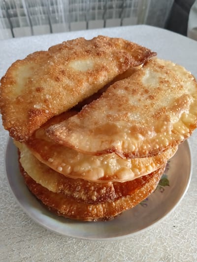 A close-up, high-angle shot shows a stack of chebureki, a savory pastry, on a plate. The chebureki are golden brown and appear to be fried, with visible bubbles and a slightly irregular, crimped edge. They are arranged in a slightly offset pile, with the top two chebureki overlapping. The plate has a subtle floral pattern. The background is blurred, suggesting an indoor setting. The surface beneath the plate has a textured, light-colored pattern. There are no people visible in the image. The scene depicts a food item likely prepared and presented in a domestic environment in Nizhnetroitskiy, Russia.
