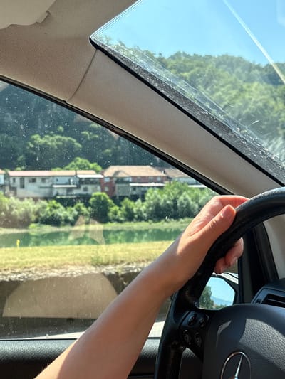 A first-person perspective shot from inside a car shows a person's hand on the steering wheel. Outside the vehicle, a body of water is visible, with a town situated on the opposite bank, nestled at the foot of a lush, green hillside under a clear blue sky. The town's buildings, featuring terracotta roofs, are clustered together. The scene appears to be along the Drina River, potentially in or near Zvornik, Bosnia and Herzegovina, given the mountainous terrain and the presence of the river. The windshield shows some dirt and water spots.