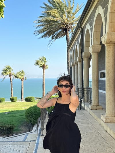 A woman stands on a paved walkway outside a stone building with arched columns. She is wearing a black spaghetti strap dress and sunglasses. Her hands are near her hair. Behind her, palm trees and green grass are visible, with a body of water in the background. The location is likely in the city of Emek HaYarden Regional Council, Israel.