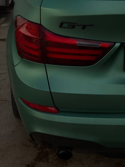 A close-up shot focuses on the rear driver's side of a green matte-finish car, potentially a GT model, judging by the "GT" emblem on the trunk. The left taillight is prominently featured, with the right side of the frame showing the car's bumper, exhaust pipe, and a side marker light. The car appears to be parked on a concrete surface, with some debris visible on the ground. The image does not provide enough information to determine a specific location within Dulovo, Bulgaria. There are no people visible in the frame.