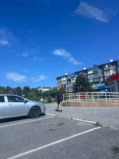 An exterior, eye-level, medium shot captures a street scene in Temryuk, Russia, under a bright blue sky dotted with wispy clouds. In the foreground to the left, a silver sedan is parked in a designated spot, its driver's side facing the viewer. The car's front fender and wheel are visible. A person, seen from the back, walks away from the car towards a building in the midground. This individual is wearing dark shorts, a dark t-shirt, and a backpack. They are walking on a paved sidewalk adjacent to a building with a colorful, multi-story facade featuring balconies with varying colored panels in blues, greens, yellows, and reds. A blue sign with white lettering that appears to read "HEBA" is affixed to the building. A white railing system is present along the sidewalk. In the background, lush green trees and other buildings are visible, including a white building with a red roof. The ground is a mix of asphalt and paved pathways.