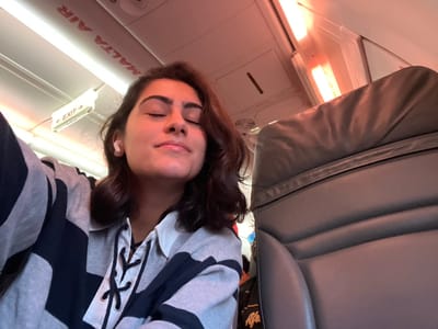 A person is lying down in a seat, possibly on an airplane, given the visible "EXIT" sign and "MALTA AIR" text. The person appears to be sleeping or resting, with their eyes closed and a relaxed expression. They are wearing a striped hoodie with black and grey patterns and white laces at the neck. The seat appears to be made of grey leather. The lighting suggests it could be daytime, but the light source seems artificial, perhaps from the cabin lights or a window. The overall mood is peaceful and relaxed. The image is taken from a low angle, looking upwards towards the person.