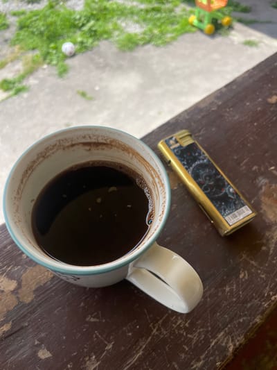 A close-up shot shows a white mug filled with dark coffee, positioned on a dark brown, weathered wooden surface. The mug has a light blue rim and some coffee residue on the inside. Next to the mug lies a gold-colored lighter with an ornate black and red design. In the background, out of focus, is a patch of green grass with a white ball and a colorful toy. The overall impression is an outdoor setting, possibly a porch or balcony, with a relaxed, casual atmosphere. The provided metadata indicates the image was taken in Alagir, Russia.